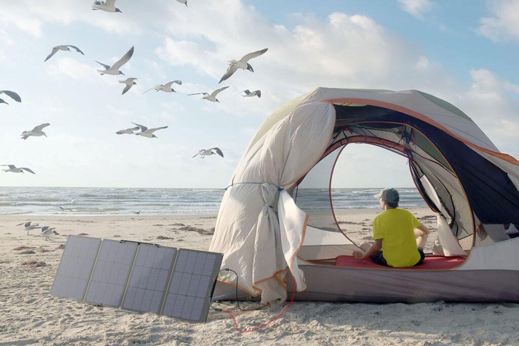 Flexibles Solarpanel von Ecoflow steht vor dem Zelt, das Zelt steht direkt am Strand vor dem Meer, im Himmel Möven und im zelt sitzt eine Frau.